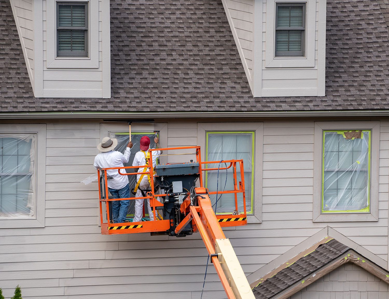 Two painting contractors on an orange bucket lift painting the second story window of a traditional home in a tan color in Charlotte, NC